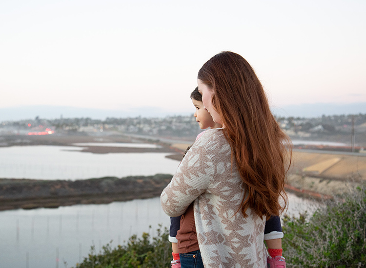 Woman holding young child in arms while they gaze at the San Elijo Lagoon and surrounding nature in San Diego.