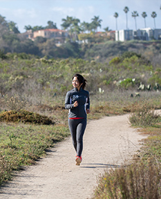 A woman is running on a dirt trail. Her surroundings show the desert coastal ecosystem that make up many San Diego hikes.