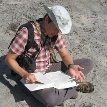 Young man with a tan hat and red plaid shirt sitting in the sand reading notes