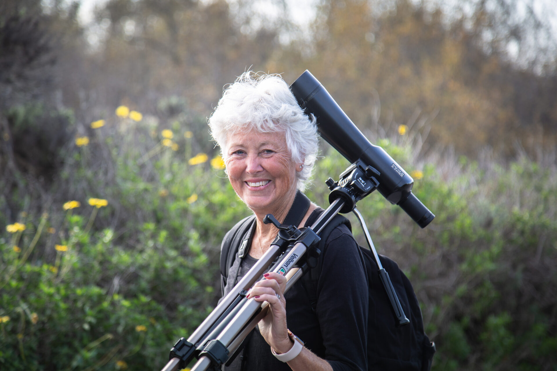 Elderly woman is smiling with a telescope in hand. Yellow flowers and other coastal wetland plants surrounding her.